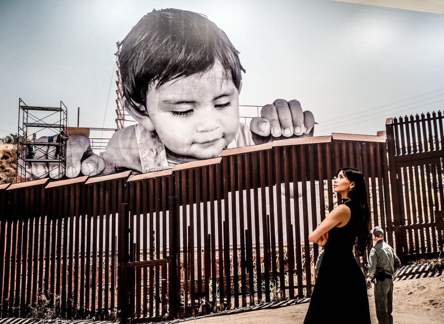 A woman stands before a large print of a photograph of artwork that had been erected atop the U.S.-Mexico border fence. In the print, a giant child appears to peer over the fence at border guards.