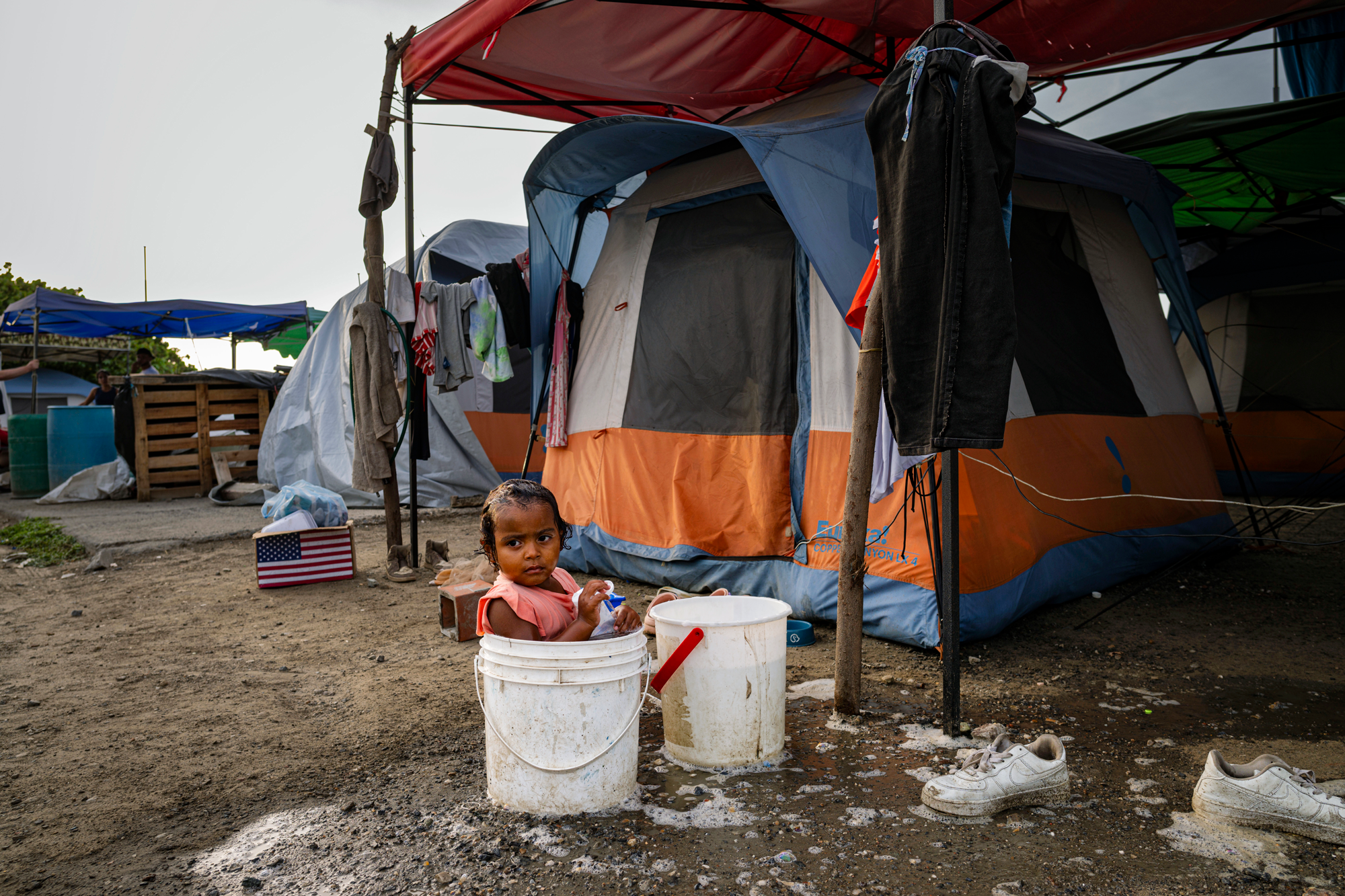 a child bathes in a bucket outside of an orange and blue tent, with an open cardboard box emblazoned with an American flag in the background