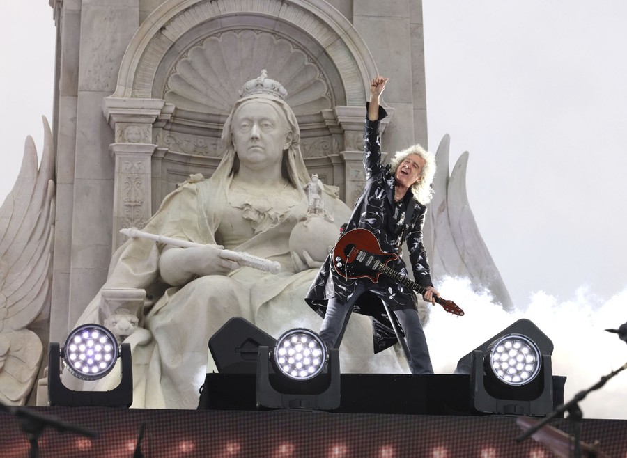 A rock star raises his arm while performing in front of a large sculpture of Queen Victoria.