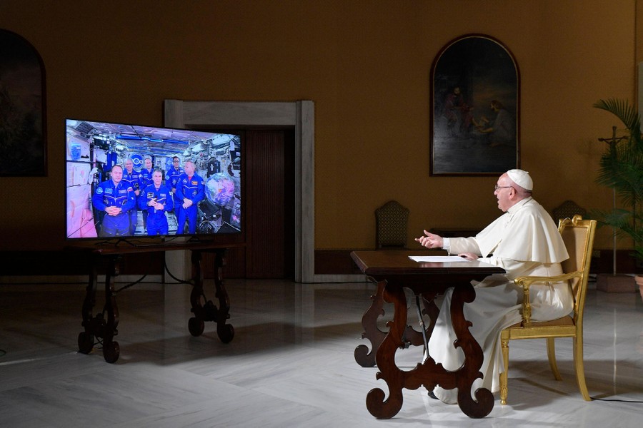 Pope Francis sits at a desk, addressing a TV screen that shows a crew aboard the International Space Station.