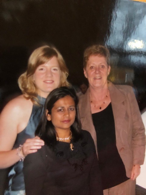 Three dressed-up women smile for the camera, one stands in front of the others.
