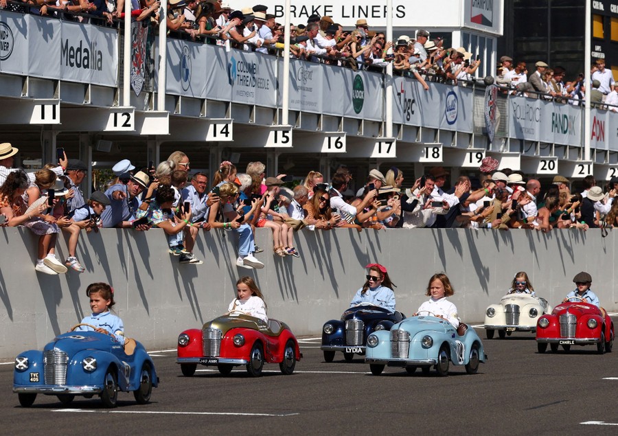Six children race pedal cars built to look like old automobiles.