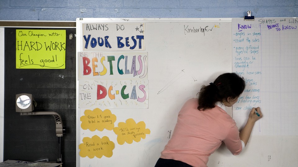 A woman writes on a dry-erase board at the front of a classroom. Her back faces the camera.