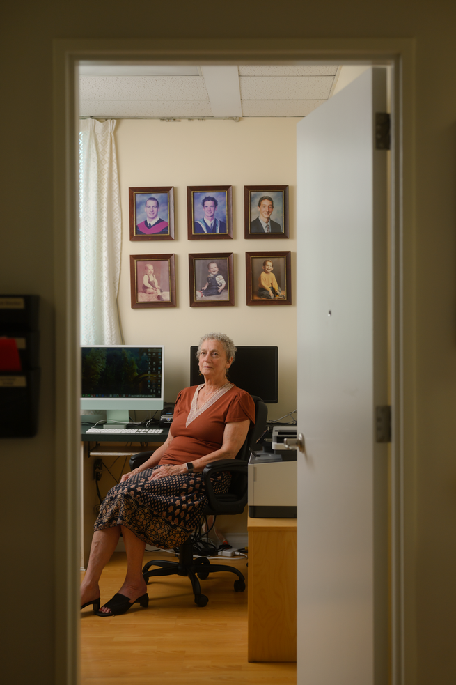 photo through open door of woman wearing brown top and patterned skirt sitting in office chair by desk with computer + 2 monitors, with 6 framed photos on wall behind