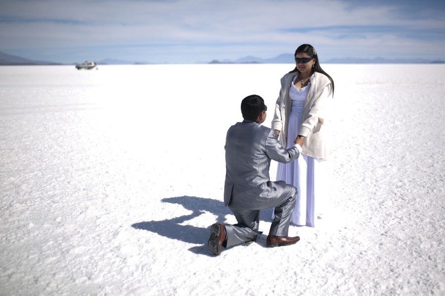 A man in a suit kneels in front of a woman wearing a white dress in the middle of a vast salt flat.