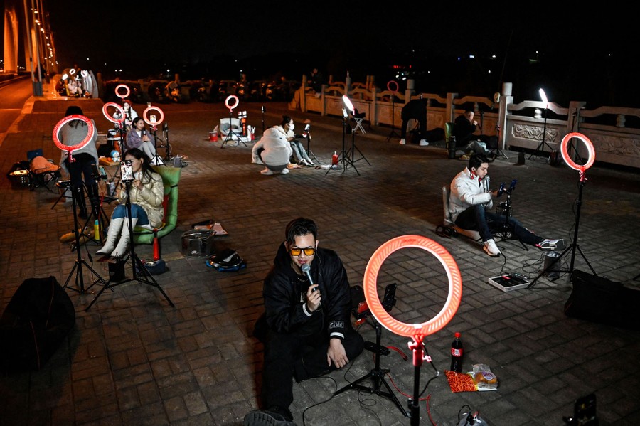 A dozen or so people sit spread out on a bridge at night, each of them with a ring light, a mobile phone on a tripod, and a microphone, livestreaming.