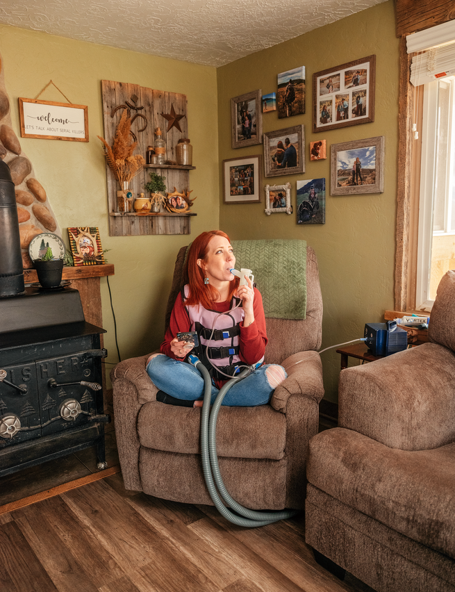 photo: red-haired woman sits cross-legged on armchair in living room inhaling from medical apparatus with long gray tubes
