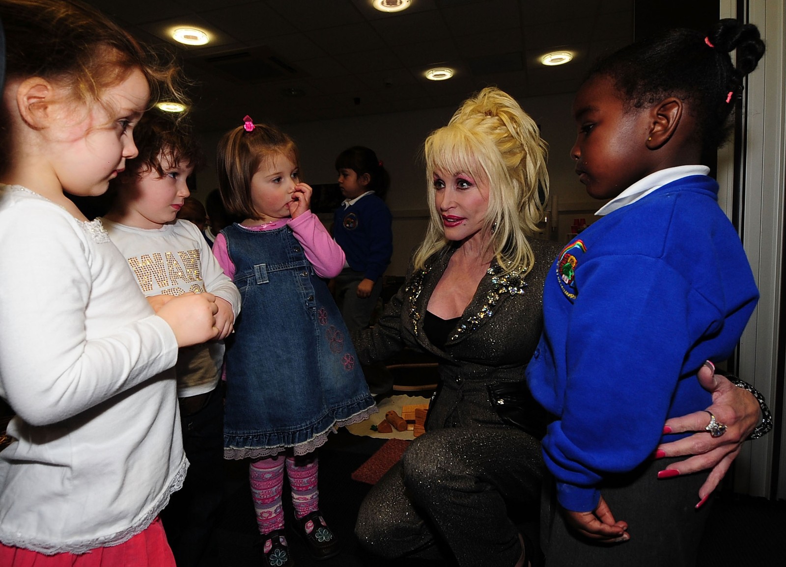 Dolly Parton kneels down, speaking with several young children.