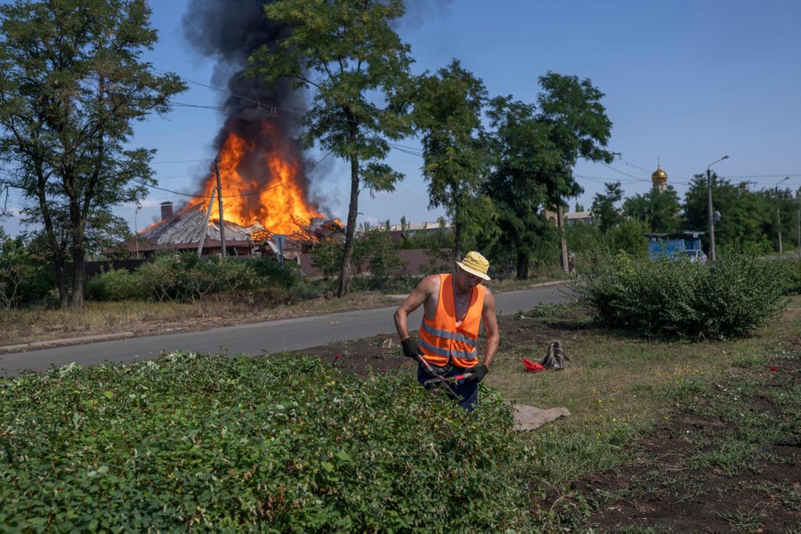 A worker trims bushes as a house burns behind them.