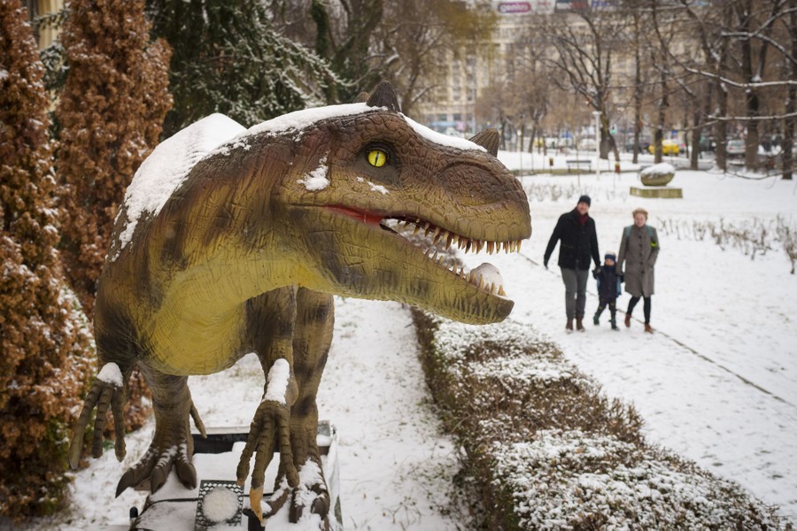 A family walks past a snow-covered model of a dinosaur.