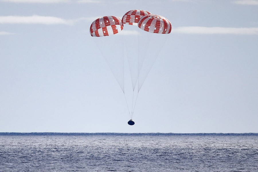 A space capsule hangs from three large parachutes, falling toward the ocean.