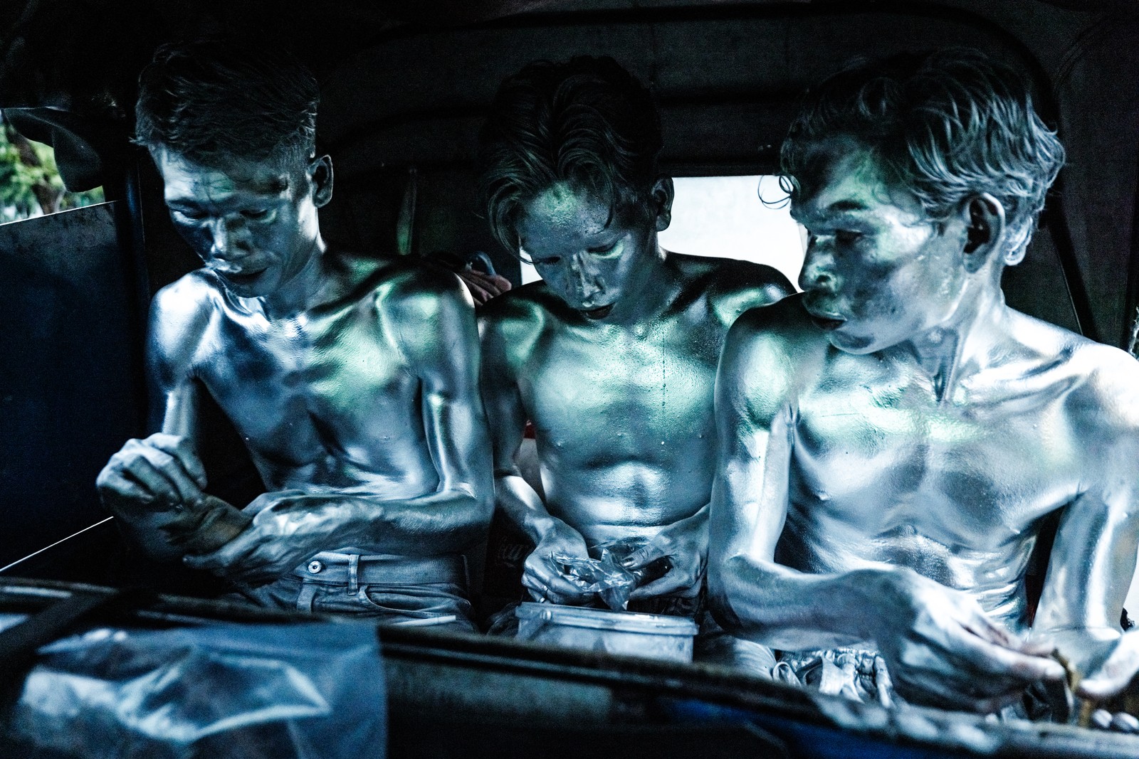Three men completely covered in silver paint sit side-by-side in a tuk-tuk, counting money.