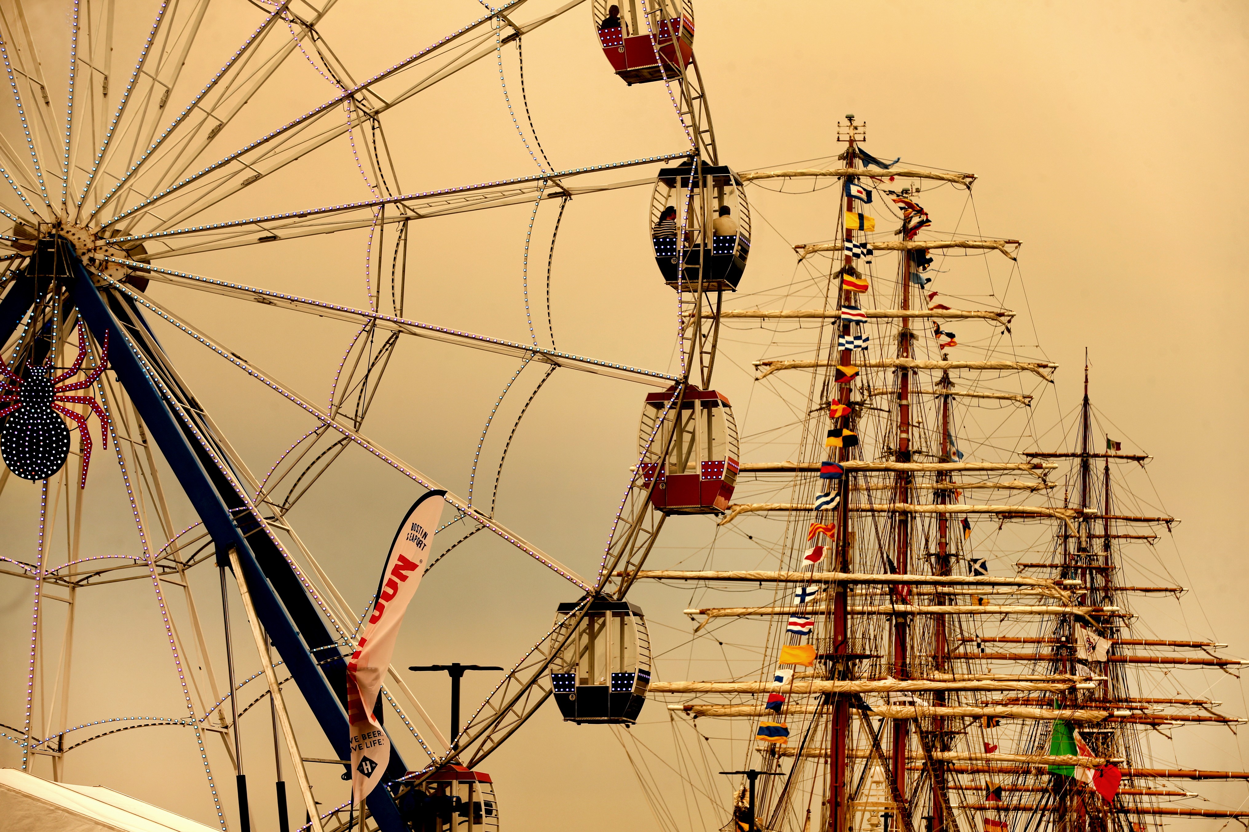 A yellowish sky forms the backdrop for a Ferris wheel and masts of at least two tall sailing ships.