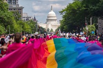 A large rainbow flag draped over a street full of parade marchers, with the U.S. Capitol Building in the background.