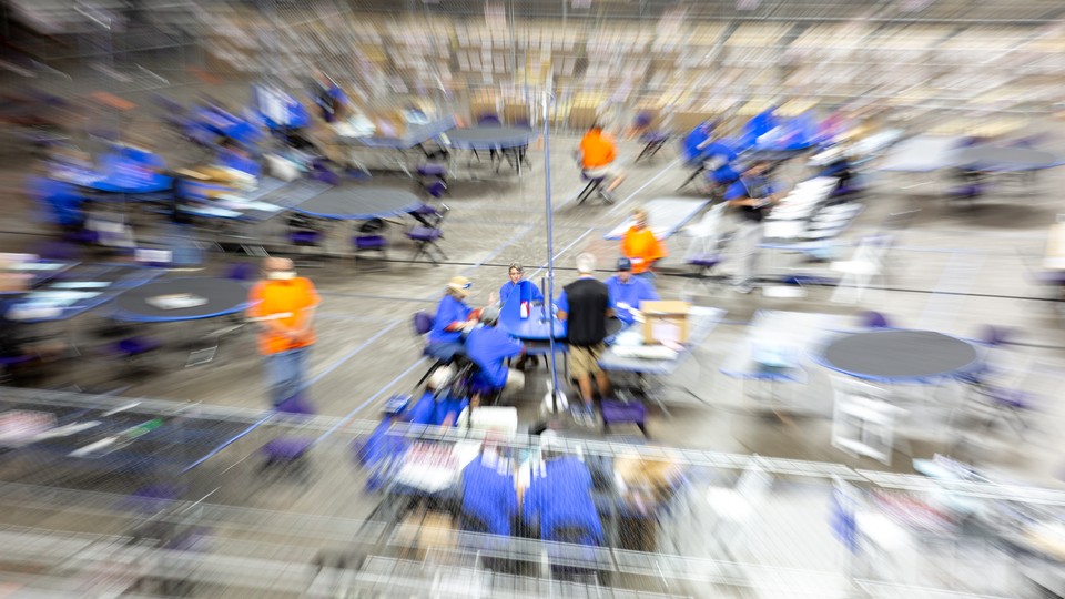 A blurred photo of groups of people in blue shirts sitting at tables during the recount in Arizona. A few people in orange shirts are dotted around the room.