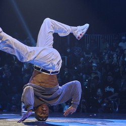 A dancer performs an acrobatic move in front of a crowd, while upside down, supported by only their head.