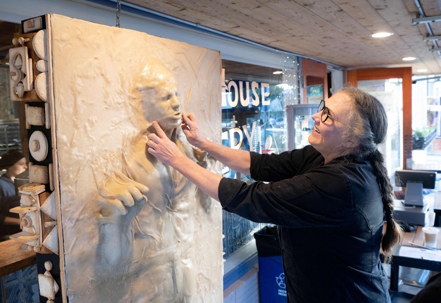 A person places their hands on a large bread-based sculpture resembling a human being frozen in a block of semisolid goo.