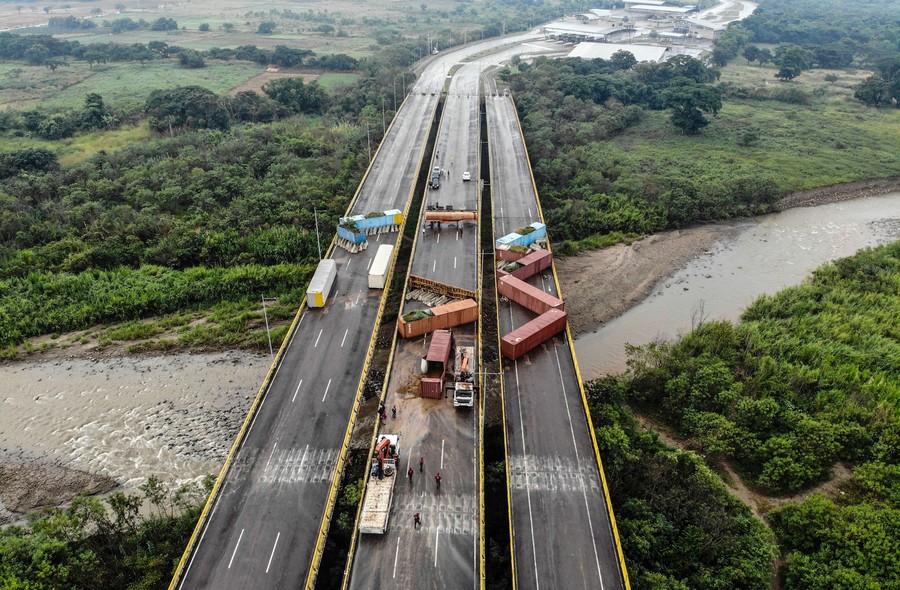 About a dozen large containers sit at extreme angles, blocking on a highway bridge over a river.