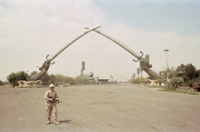 an image of an American soldier, clad in light brown fatigues and holding a rifle, standing on a road in Baghdad
