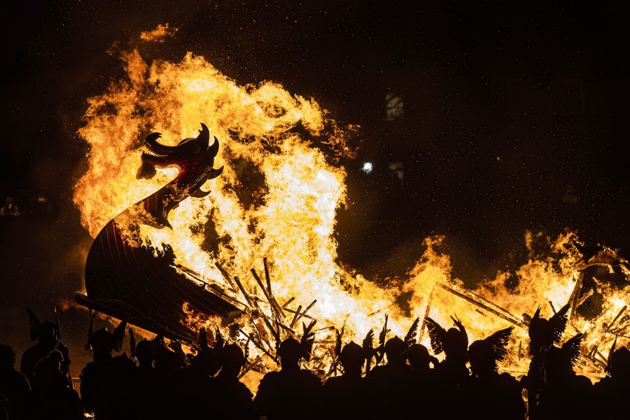Men wearing Viking costumes are silhouetted against flames from a burning longboat.