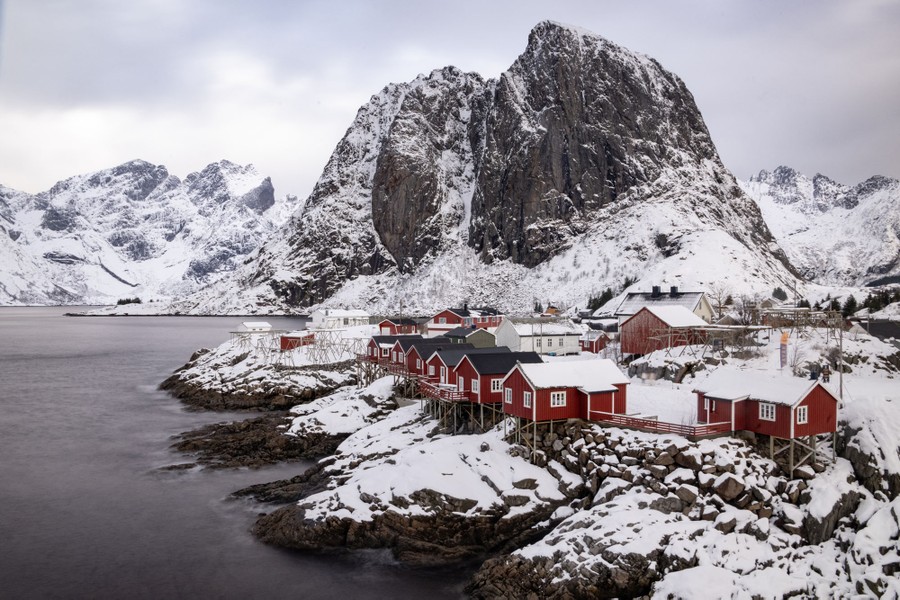 A picturesque fishing village on a rocky shoreline, backdropped by dramatic, steep mountains, all covered in a dusting of snow.