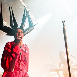 A performer onstage wearing a large, spiky headpiece made of plastic