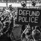 black-and-white photo of protest crowd with fists raised, with sign "DEFUND POLICE"