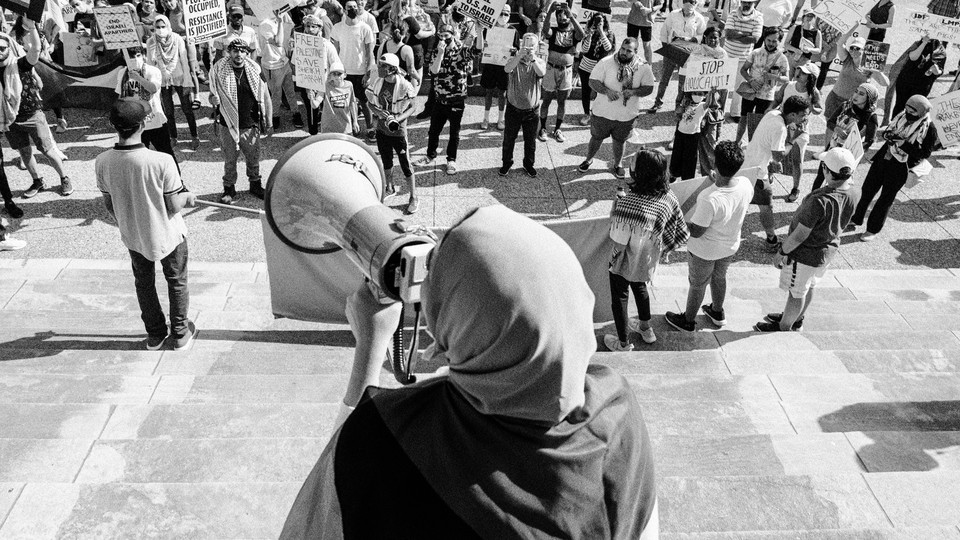 Picture showing an activist wearing a hijab holding a megaphone, speaking to a crowd of protesters holding signs. One of the signs reads: "Free Palestine."