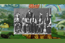 A black-and-white posed photograph of seated members of the Red Cloud delegation adorned in traditional regalia. Behind them stands a man in a suit.