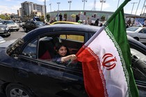 Photograph of a young girl sitting in the backseat of a car and holding an Iranian flag out of the rolled down window