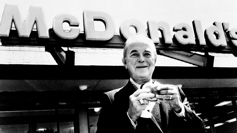 Ray Kroc eats a hamburger in front of a McDonald's sign