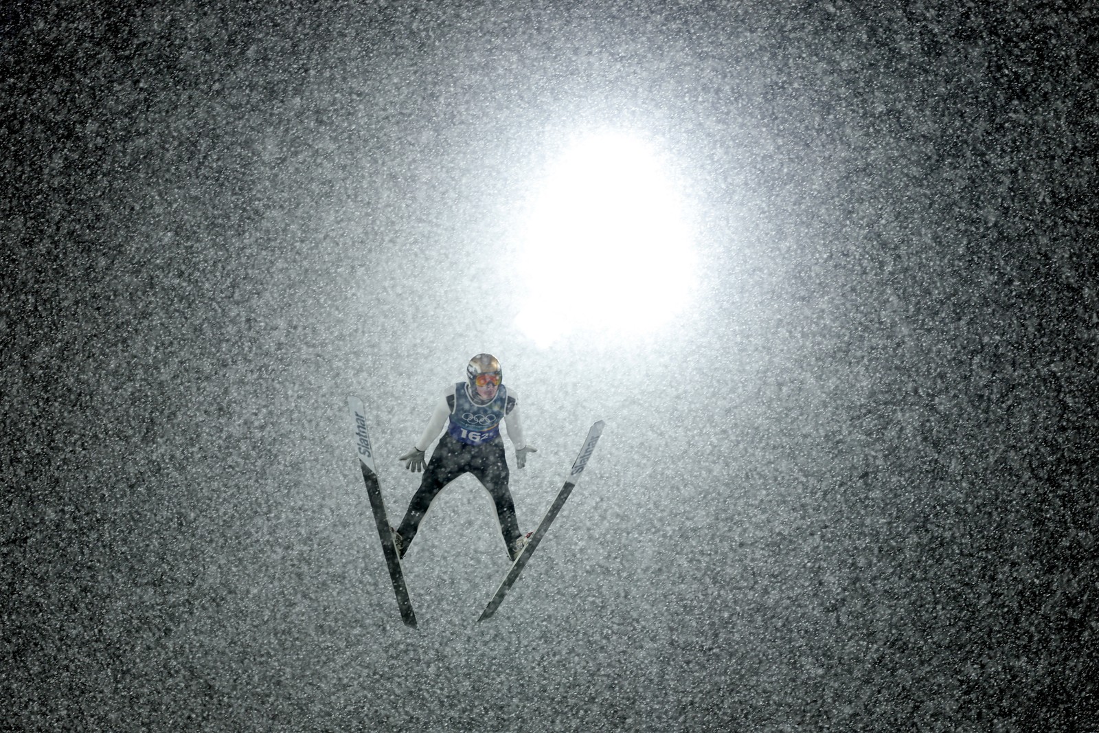 A ski jumper flies through a flurry of snowflakes at night, backlit by a bright light.