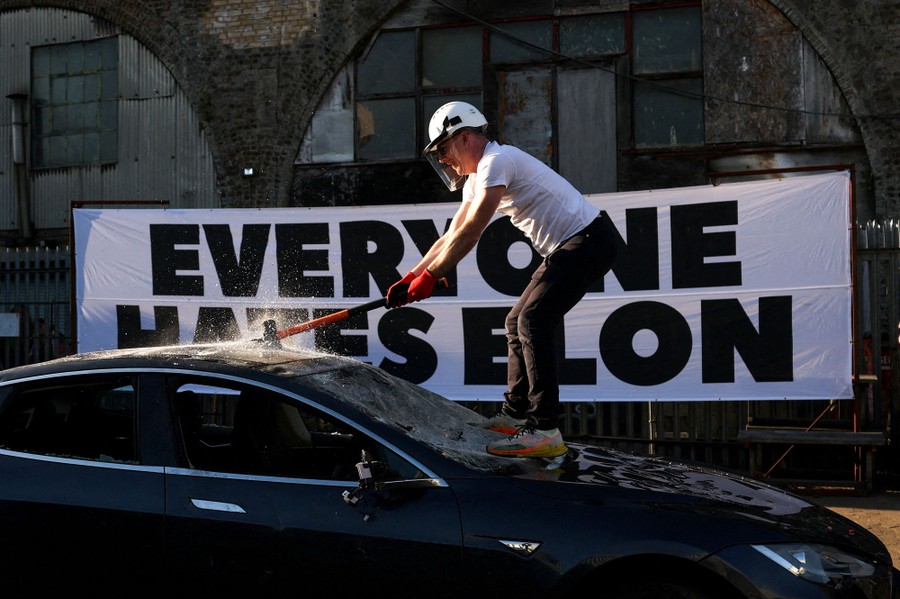 A person stands on the hood of a Tesla car, swinging a sledgehammer at its roof, standing in front of a large banner that reads "Everyone hates Elon."