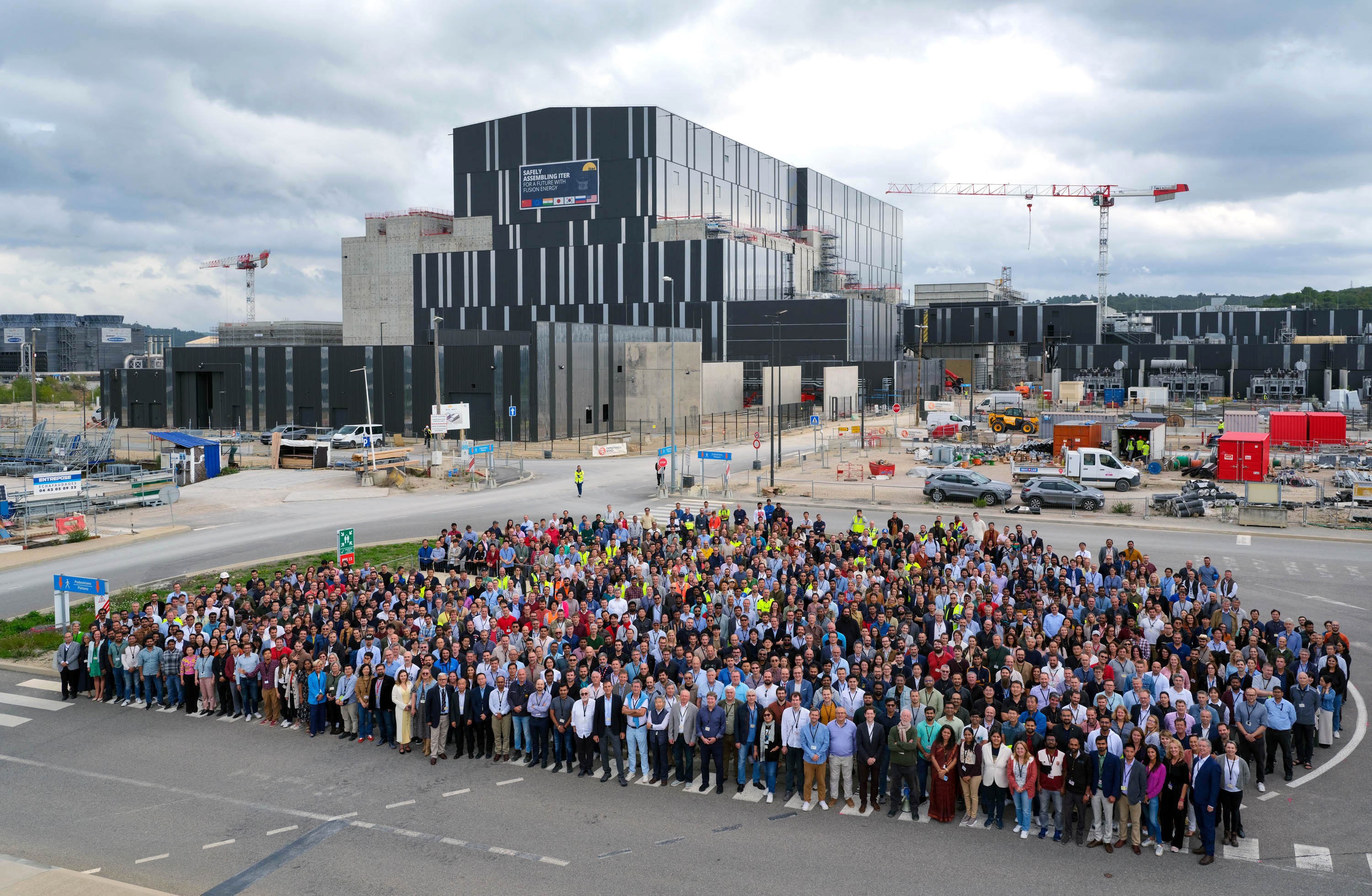 Hundreds of people gather beside a road for a group photo in front of a large facility.