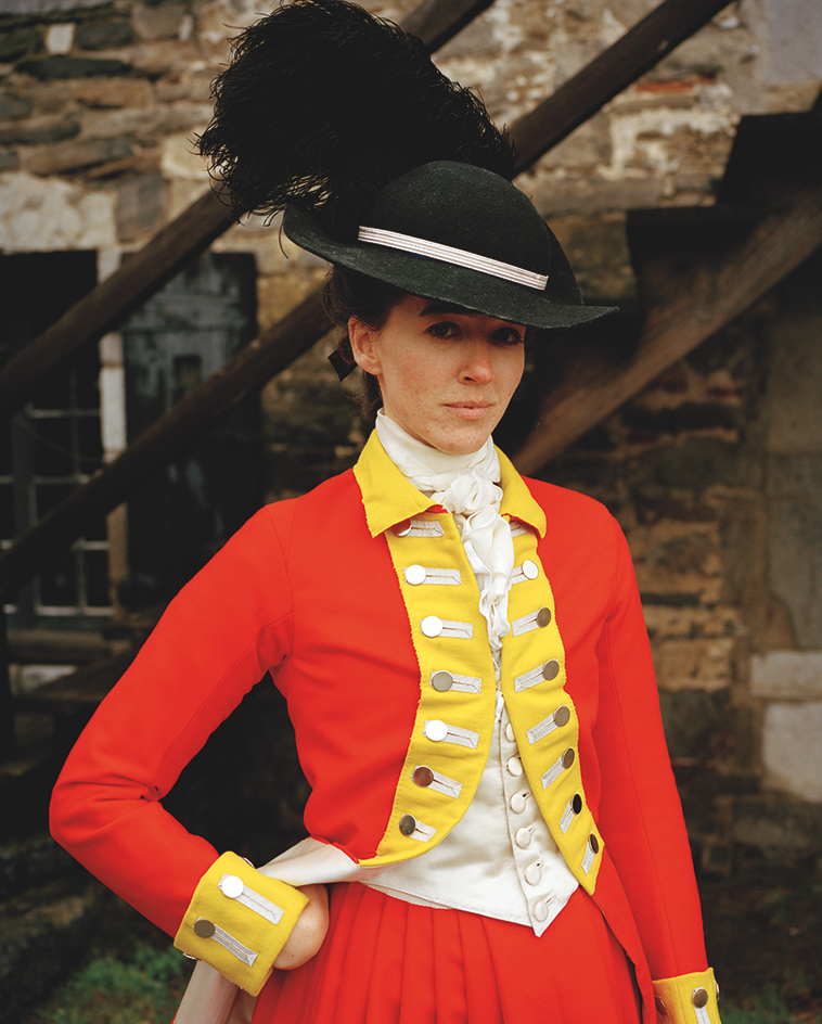 photo of a female reenactor as a British soldier wearing a large plumed black hat, orange-red coat and trousers with yellow lapels and cuffs, standing by wooden stairs with with one hand on hip, inside the grounds of Fort Ticonderoga