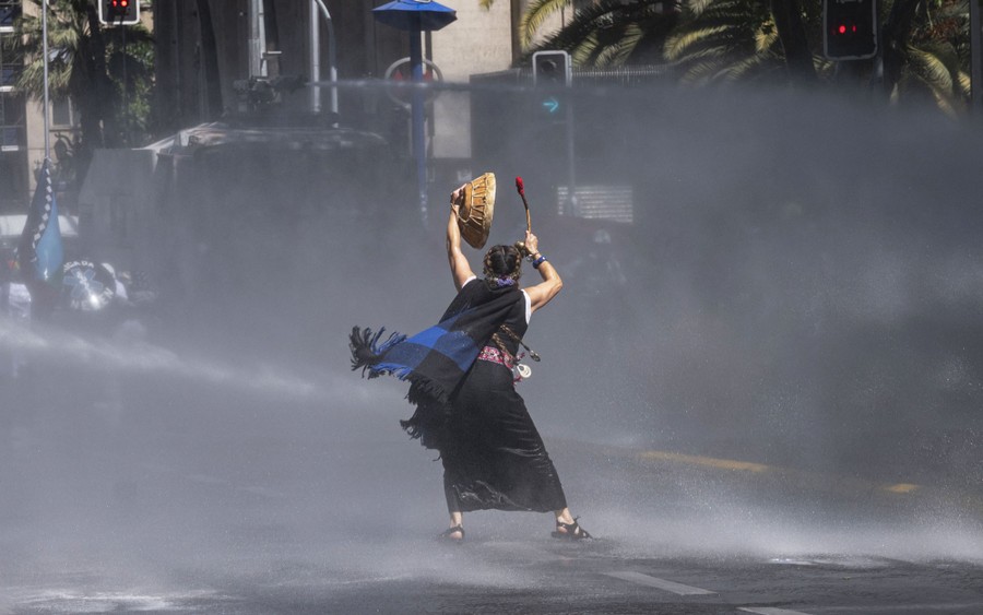 A person stands in a street during a protest and is sprayed with water from a water cannon.
