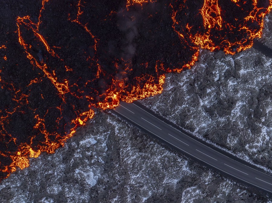 An aerial view of lava flowing over a road and rocky ground dusted with snow