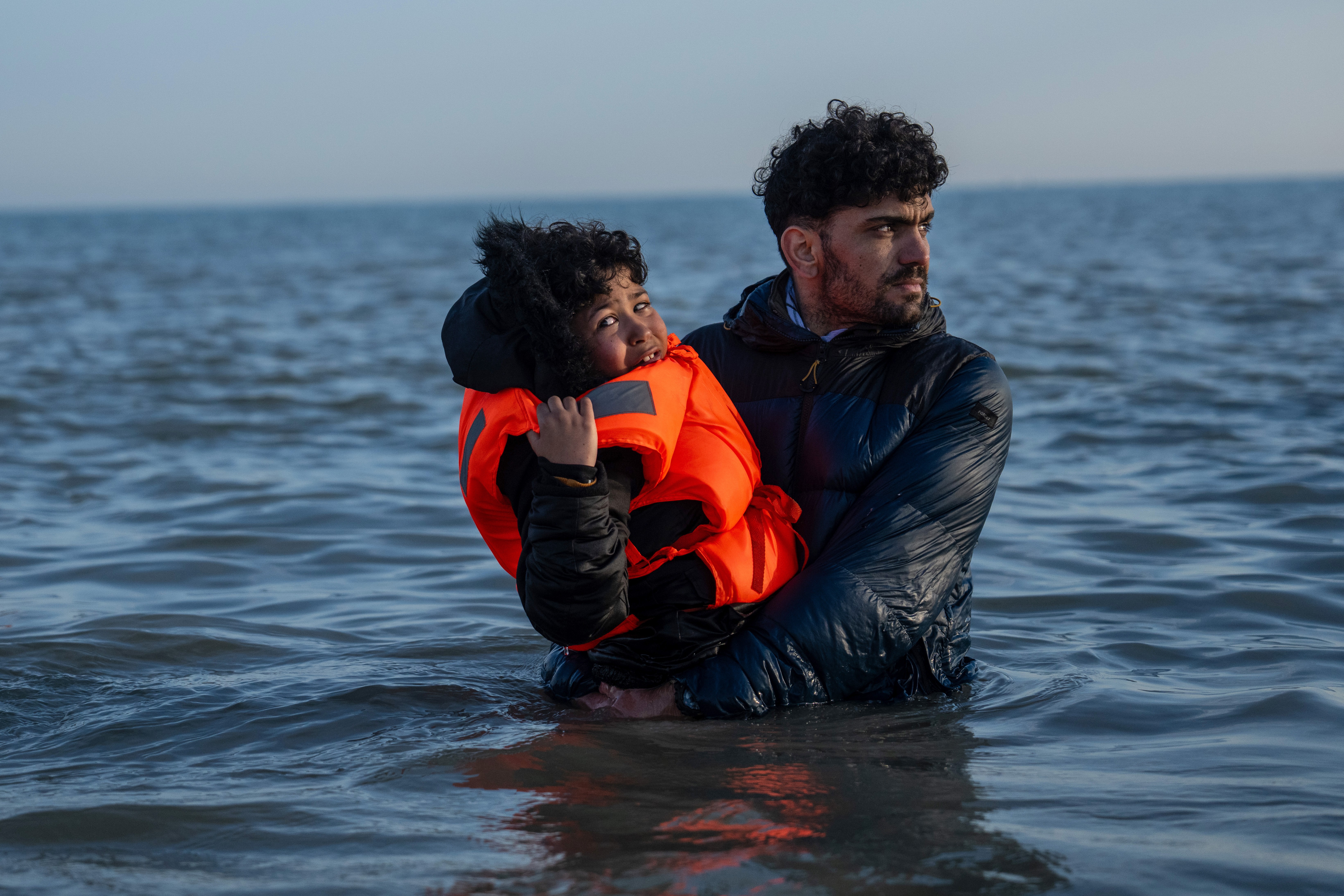 A boy wearing a life preserver is carried by a man through waist-deep water.