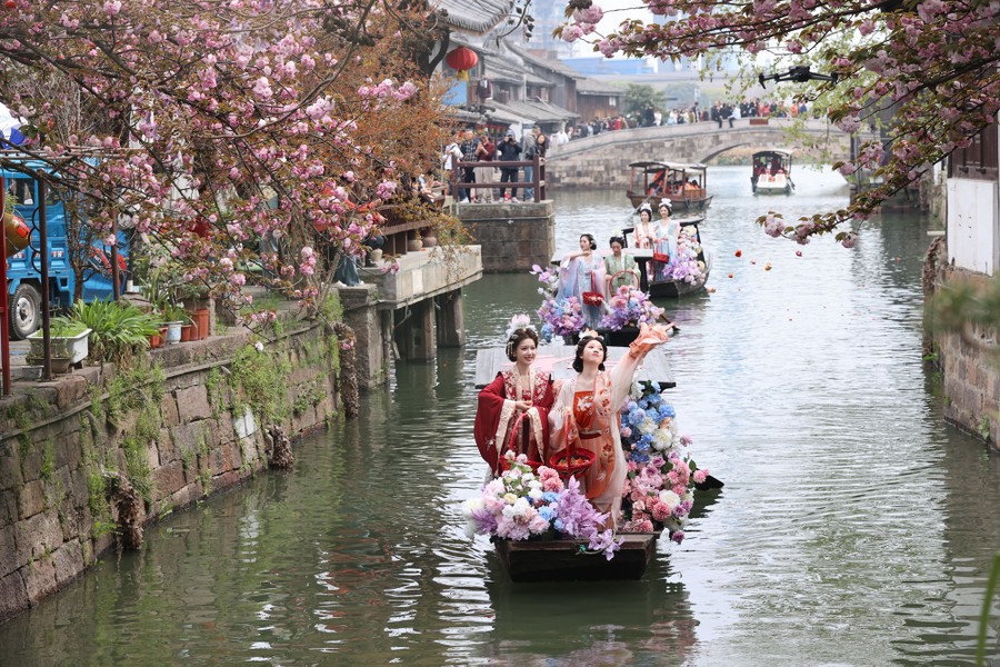 Women in traditional costumes stand in small boats, tossing flower petals as they navigate through a canal during a festival.