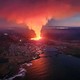 An aerial view of a coastal Icelandic town with an erupting volcanic fissure a short distance inland, just uphill from its houses