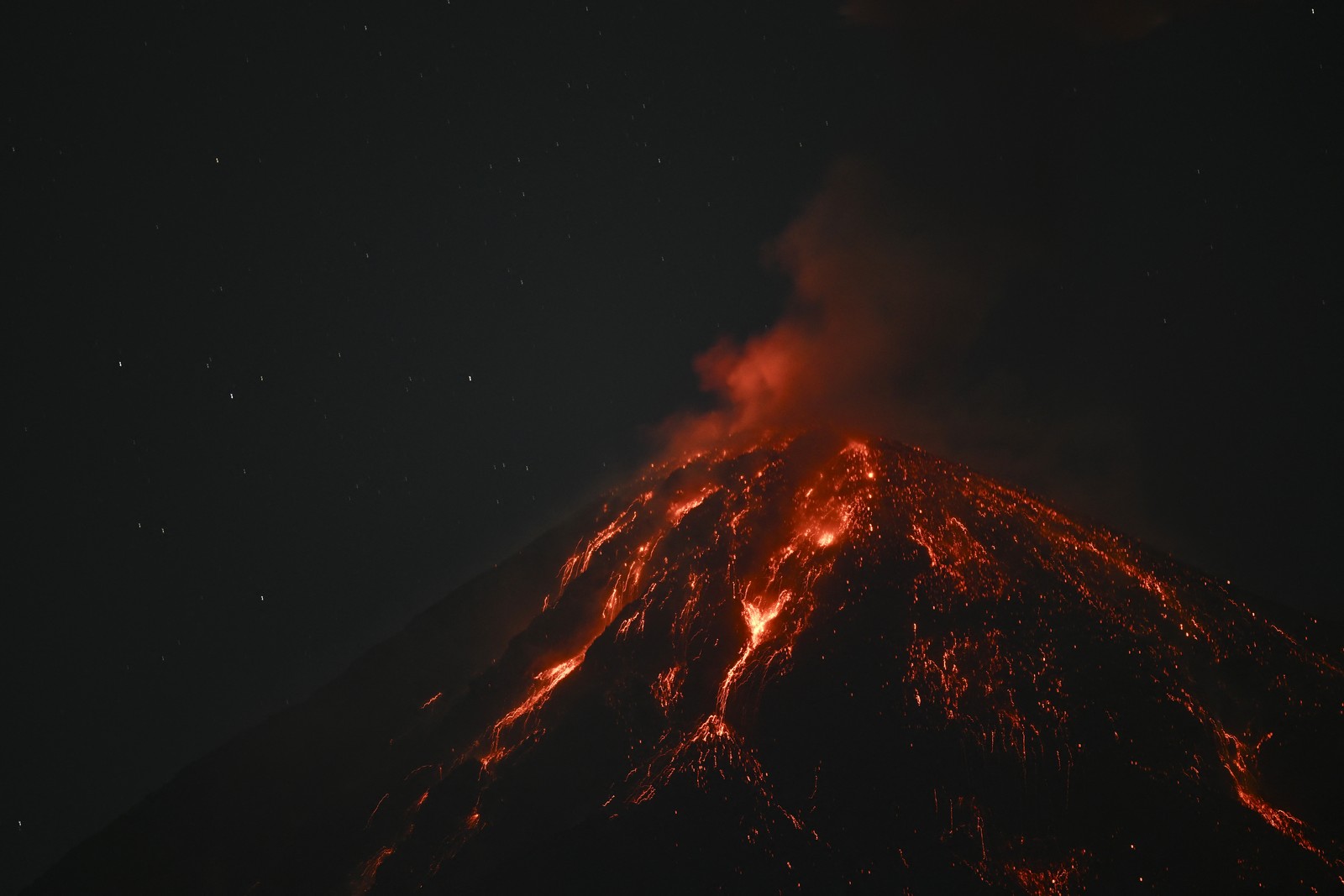 Lava glows across the top of an erupting volcano, seen at night.
