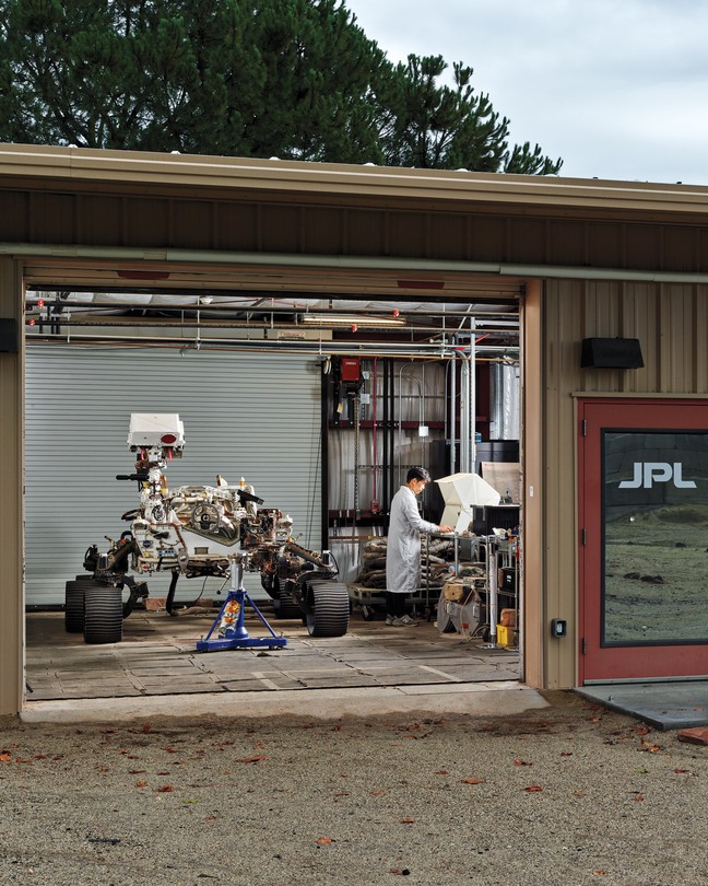 Color photo of one-story building with garage door open to reveal a wheeled rover and a person in white coat working at a bench. A sign on the building reads "Mars Yard."
