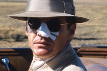 A movie still of Jack Nicholson, playing private eye J. J. Gittes in Chinatown (1974), in a convertible car with the top down, wearing a fedora and sunglasses with his nose covered in white medical tape.