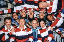 group photo of 10 astronauts in space station, some upside down, most wearing matching red, white, and blue polo shirts