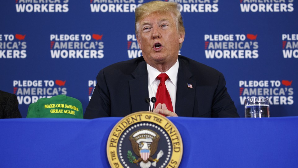 President Donald Trump sits at a blue table with a "Make Our Farmers Great Again" hat