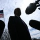 An image of Trump's silhouette from below surrounded by microphones, with an American flag flying in the background.