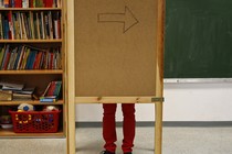 A pair of legs are visible behind a wooden corkboard in a classroom. 