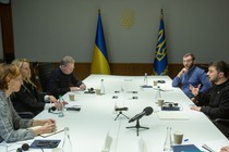 Several people sit around a large table framed by two blue-and-yellow flags; Volodymyr Zelensky speaks while several others take notes.