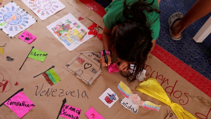 A girl in a green shirt draws hearts and houses on a paper labeled "Venezuela."