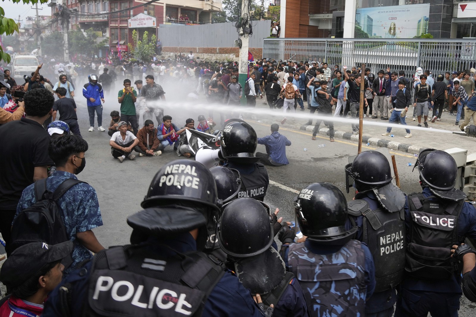 Riot police spray water from a hose at protesters who are sitting in a street.
